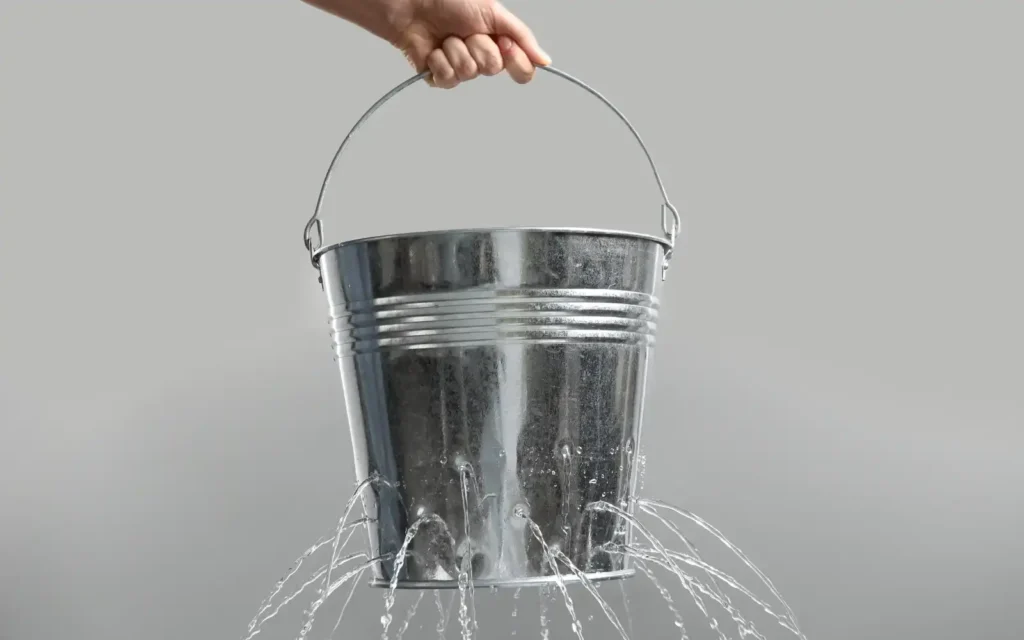 Leaking-client-funnel A metal bucket with holes in the bottom leaking water, representing a leaking client acquisition funnel