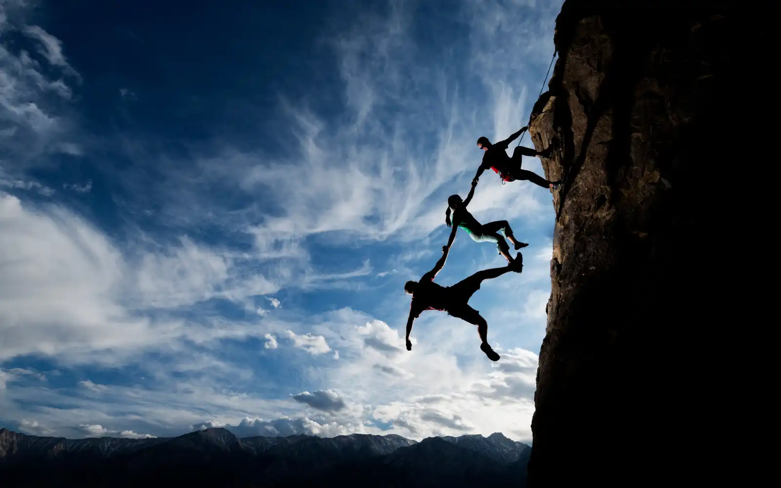 Three people sillhouetted dangling off a cliff face and holding onto each other by their hands, representing trust